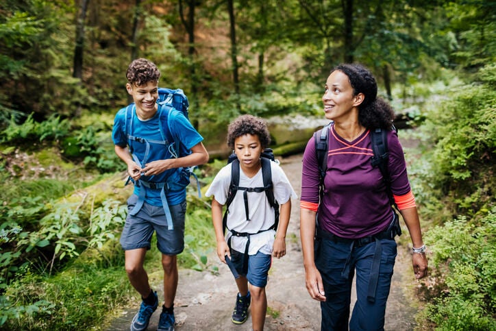 Women with her two children wearing athletic clothing and hiking backpacks smiling while hiking on a woodland trail.