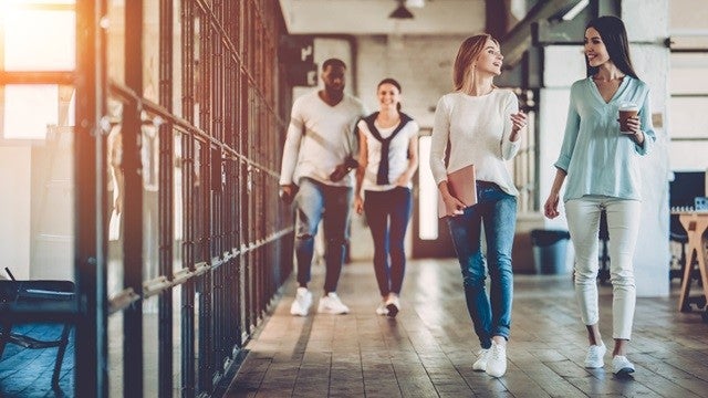 Four people walking down a modern office hallway, casually talking and smiling, with sunlight streaming through large windows.