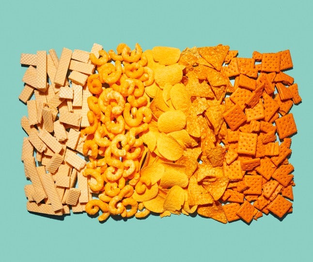 Assorted processed snacks arranged in rows on a light teal background, including wafers, cheese curls, potato chips, tortilla chips, and cheese crackers.