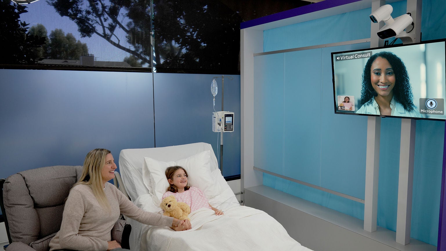 Child in a hospital bed and her mother speaking with a clinician on a wall-mounted virtual consult screen