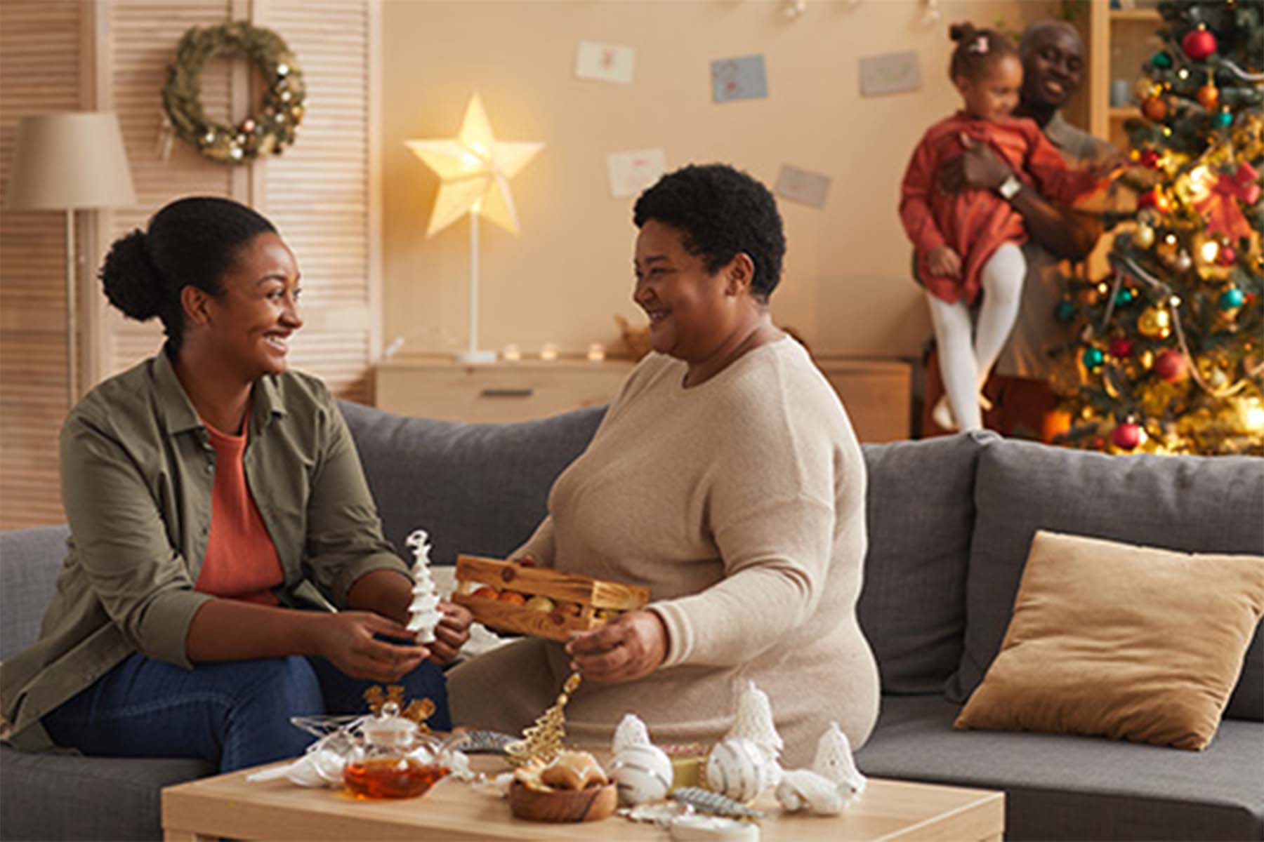 Two women decorating a living room for Christmas, with a man holding a child near a Christmas tree in the background.