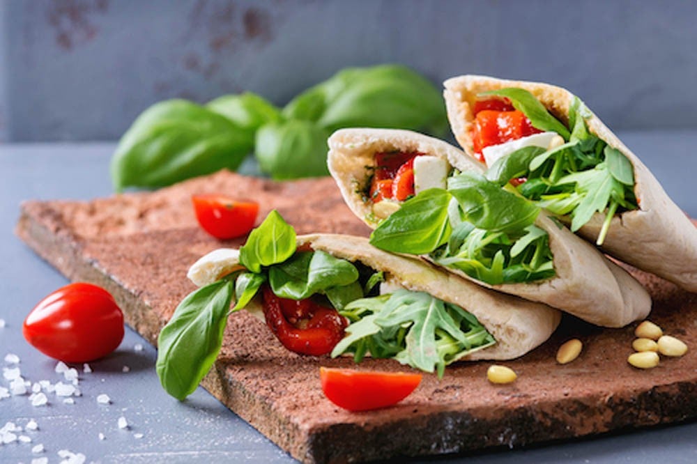Sándwiches de pan pita con verduras a la parrilla: pimiento, berenjena, tomate, albahaca y queso feta, servidos sobre una tabla de terracota sobre fondo de piedra gris