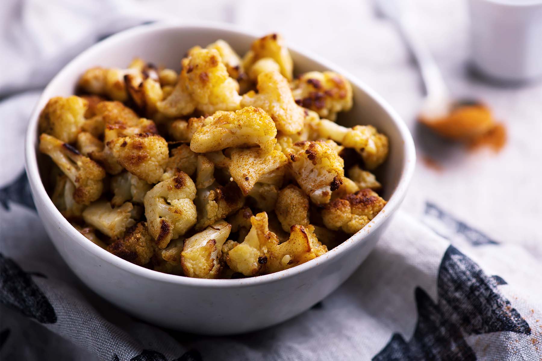 A bowl filled with roasted cauliflower florets, golden and crispy, placed on a textured cloth with a subtle pattern in the background.