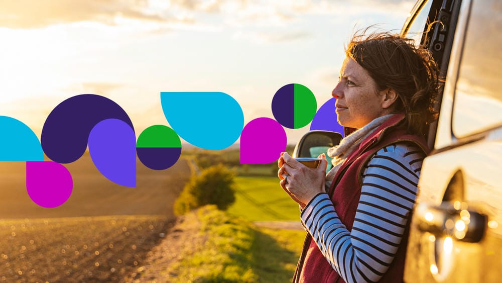 Woman standing beside a vehicle, holding a cup, overlooking fields at sunset with vibrant purple, blue, and cyan shapes in the foreground.