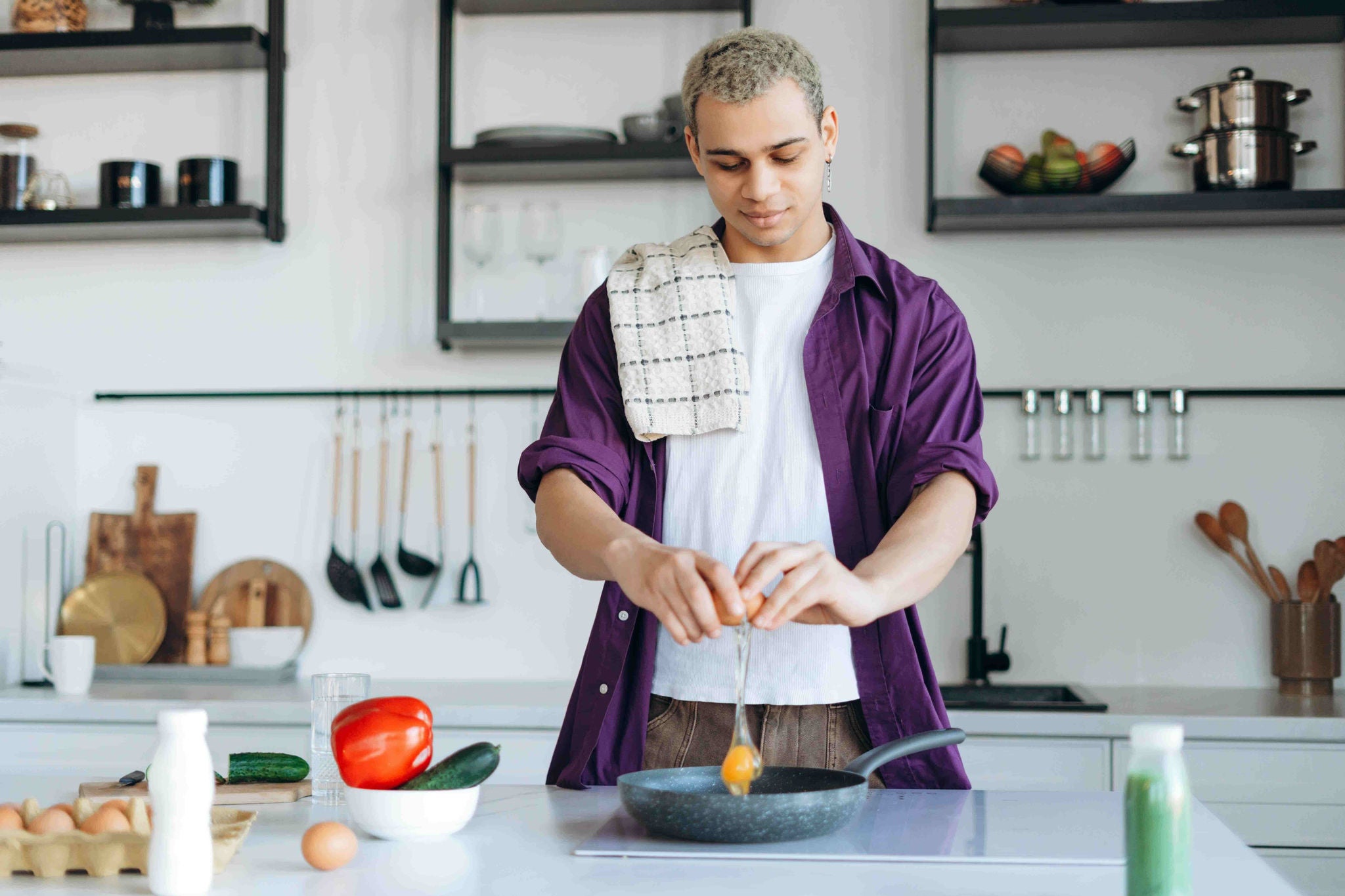 Young man cracking an egg into a frying pan in a brightly lit kitchen.