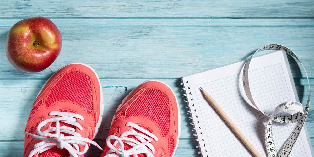Red sneakers, a red apple, a notebook with a pencil, and a measuring tape on a blue wooden surface.