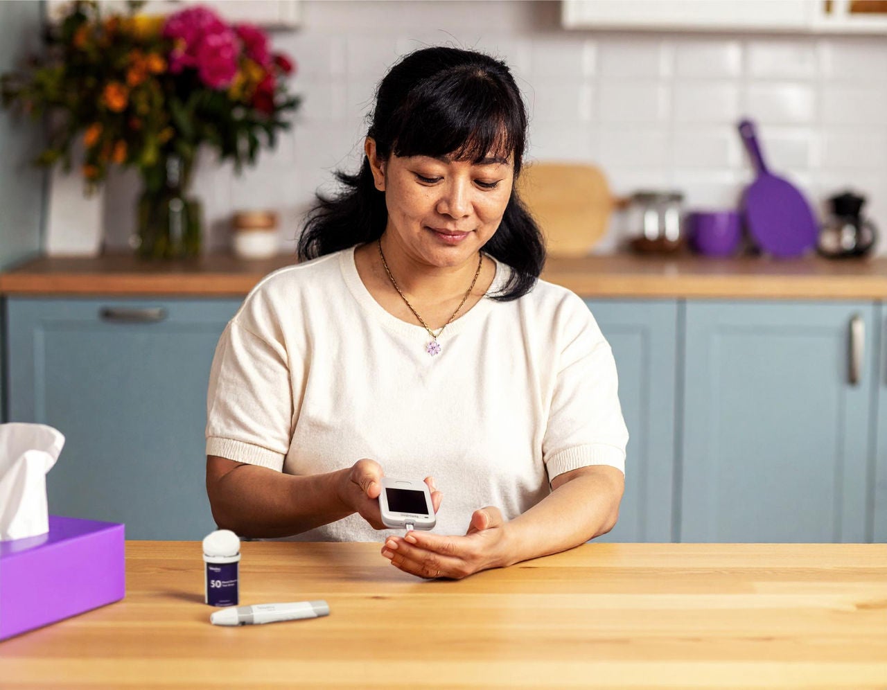 A woman checking her blood sugar using a glucose meter at a kitchen table, with test strips and a lancet nearby.
