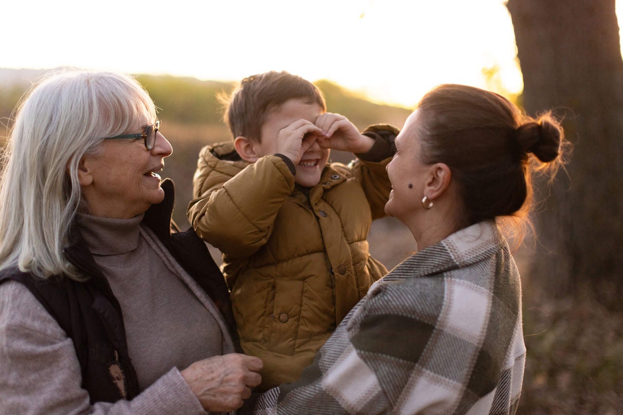 Woman holding young child with grandparent, smiling outdoors at sunset.