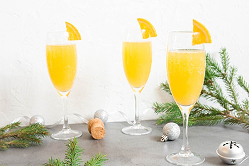 Copas de cóctel Mimosa decoradas con rodajas de naranja, rodeadas de ramas de pino y adornos navideños sobre una superficie gris
