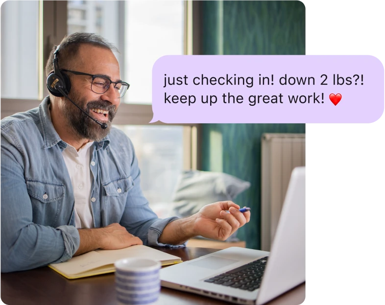 Man with a headset smiling and talking to his computer, with an overlay of a coaching message reading 'Just checking in! Down 2 lbs?! Keep up the great work!'"