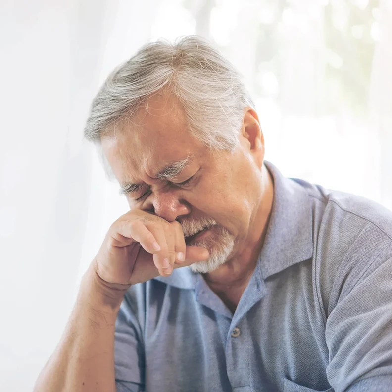 Older man sneezing into a tissue while experiencing allergy symptoms at home.