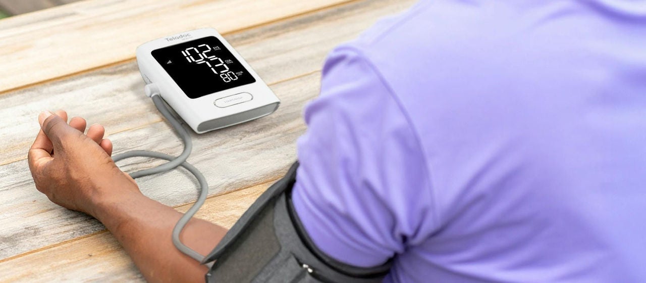 Person using a digital blood pressure monitor with an arm cuff, showing a reading of 102/75 mmHg.