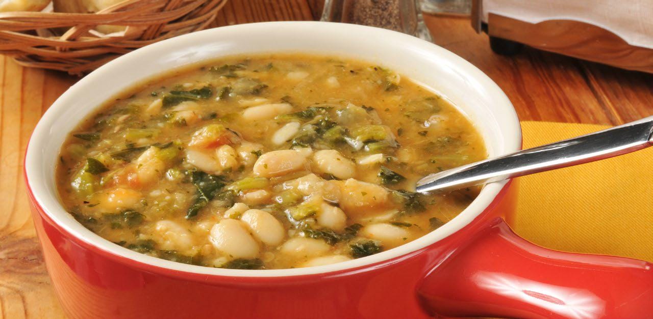 Hearty white bean and vegetable soup served in a red bowl with a spoon on a wooden table.