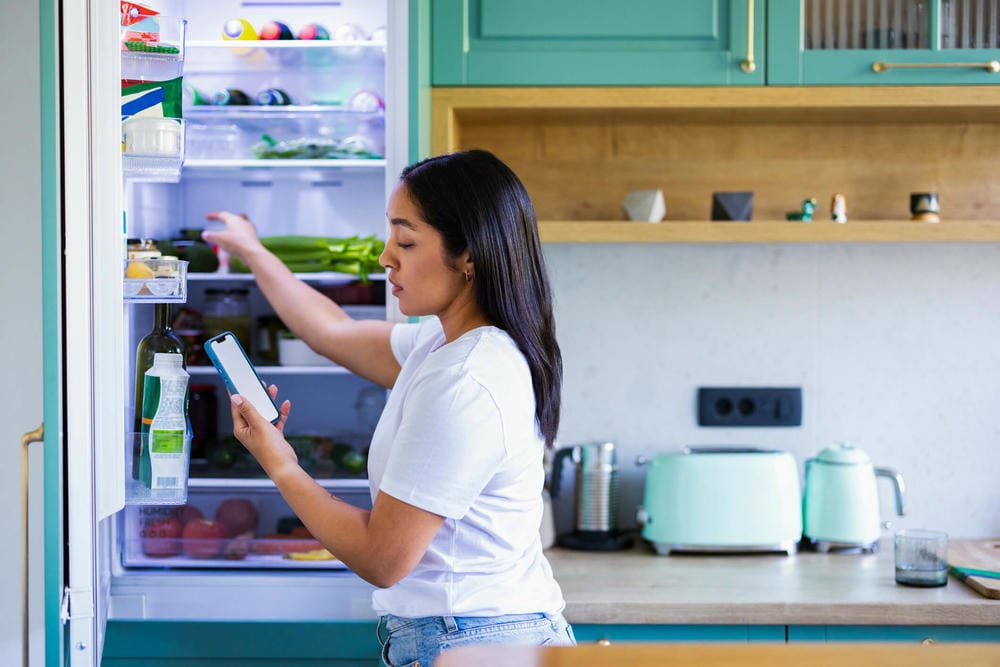 Woman looking inside an open refrigerator while holding a smartphone