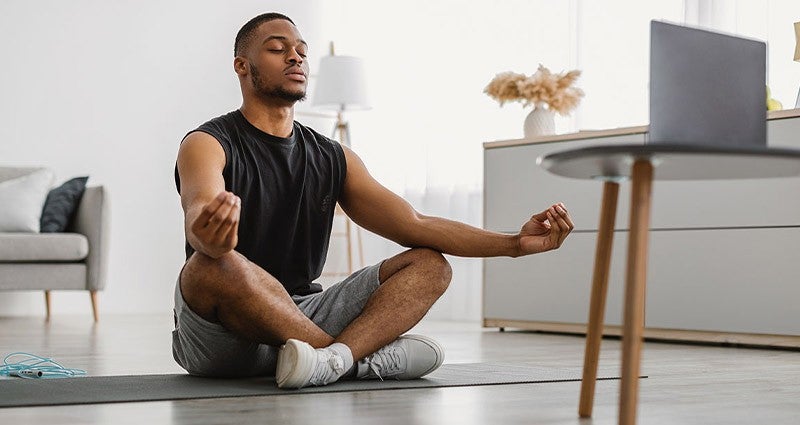 Man sitting cross-legged on a yoga mat, meditating in front of a laptop.