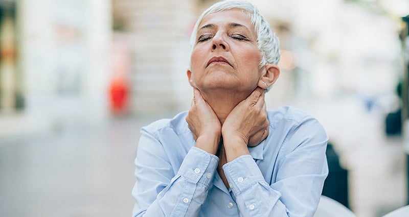 An older woman with short gray hair is sitting outdoors, looking upward with a thoughtful expression, her hands gently resting on her neck, conveying a moment of reflection.