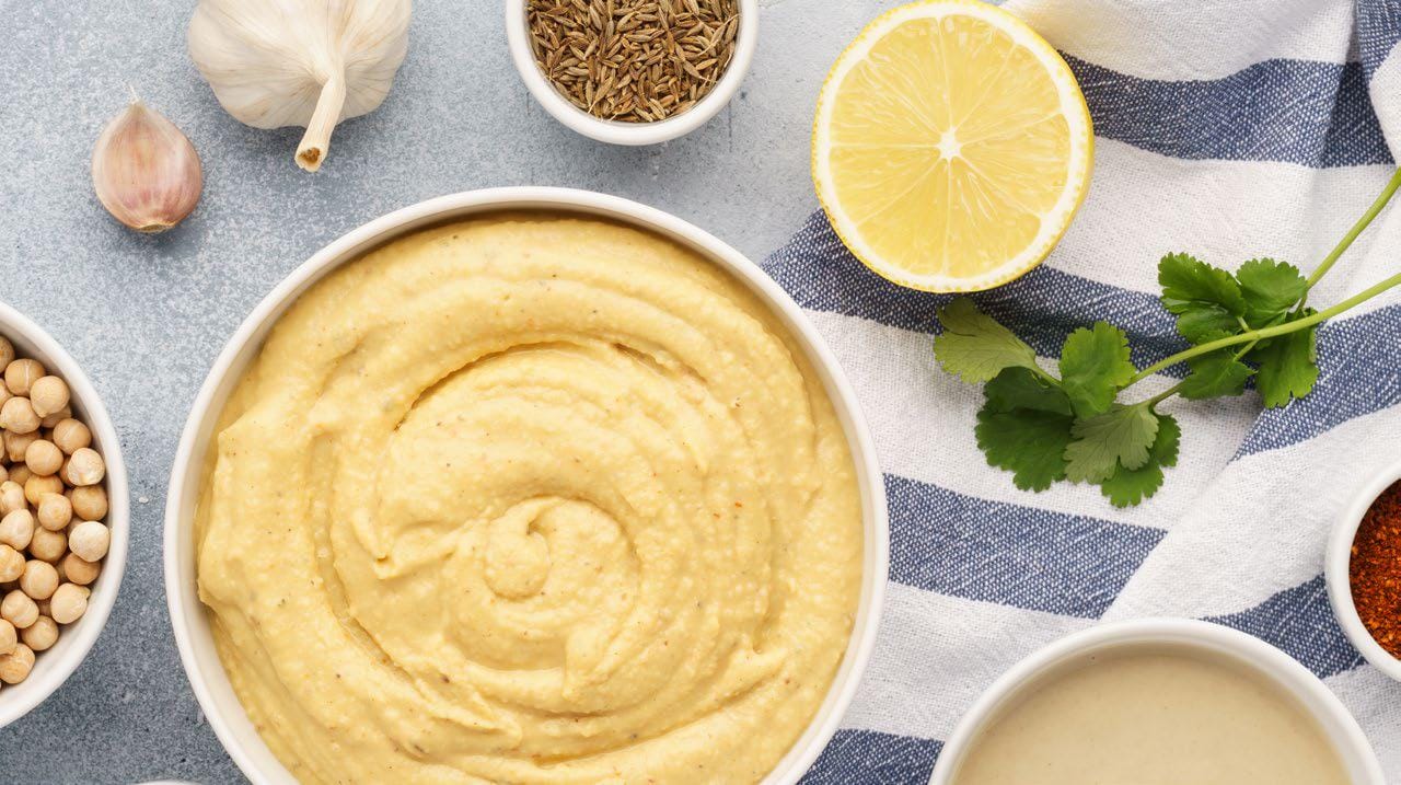 Bowl of smooth homemade hummus surrounded by chickpeas, garlic, lemon, herbs, and spices on a kitchen surface.
