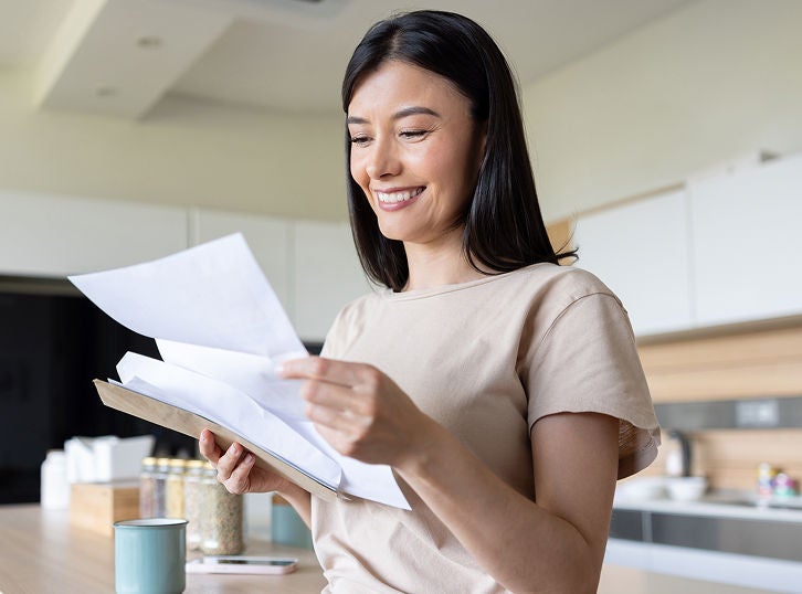 Woman standing in a modern kitchen, smiling while reviewing documents and holding a notebook.