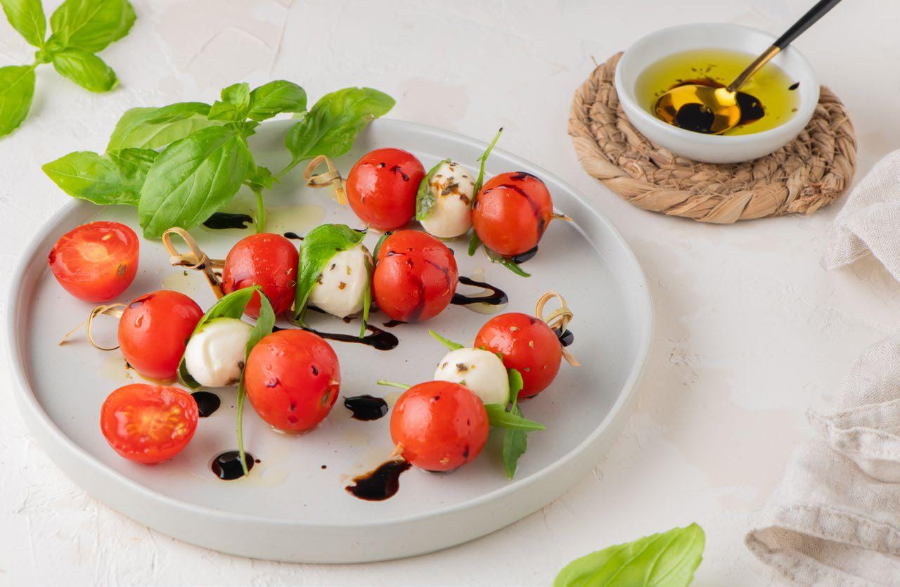 Caprese skewers on a plate with basil leaves and a small bowl with olive oil with a spoon in it
