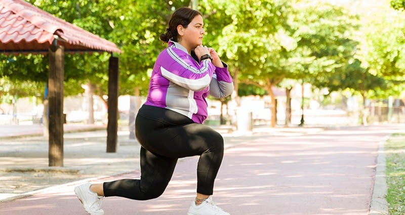 A woman is exercising outdoors, performing a lunge on a pathway surrounded by trees. She is wearing a purple and white top with black leggings, indicating an active lifestyle.