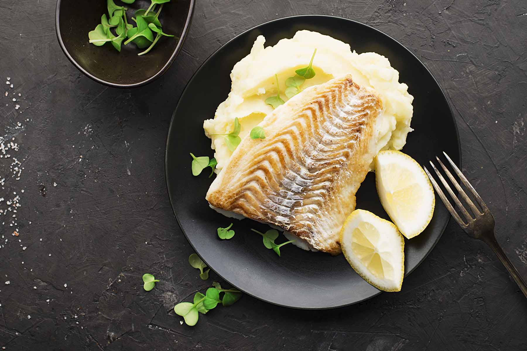 A plate featuring a portion of grilled fish with lemon wedges, served alongside creamy mashed potatoes and garnished with fresh herbs, on a dark textured surface.