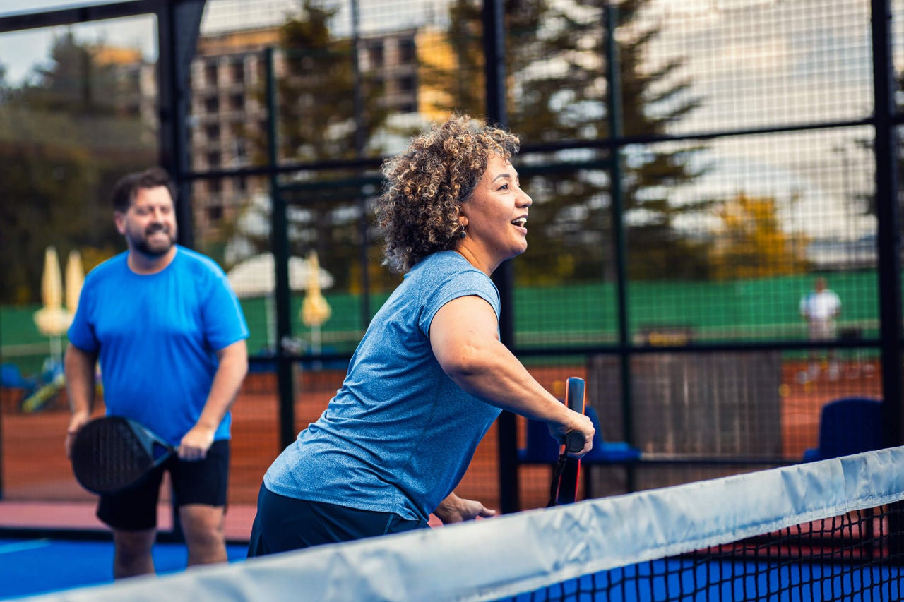 Man and woman wearing athletic clothing playing pickleball on an outdoor court while smiling. 