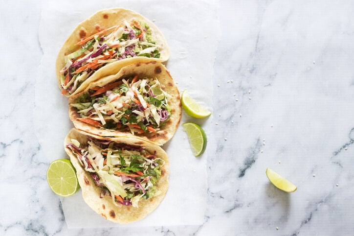 Three tacos with shredded cabbage, carrots, and fish garnished with limes on a marble surface.