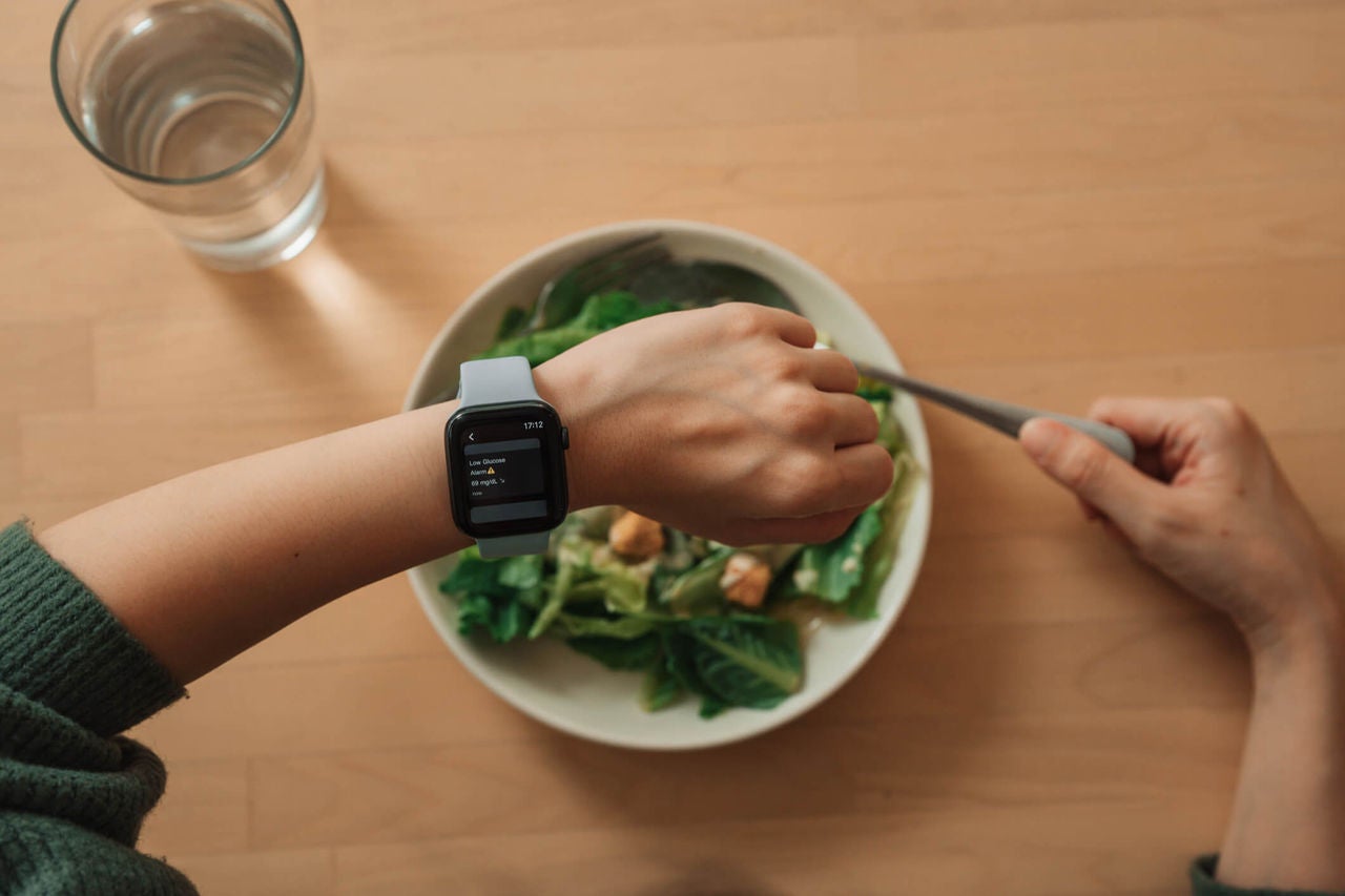 Person checking smart watch with blood sugar tracking information over a bowl of salad.