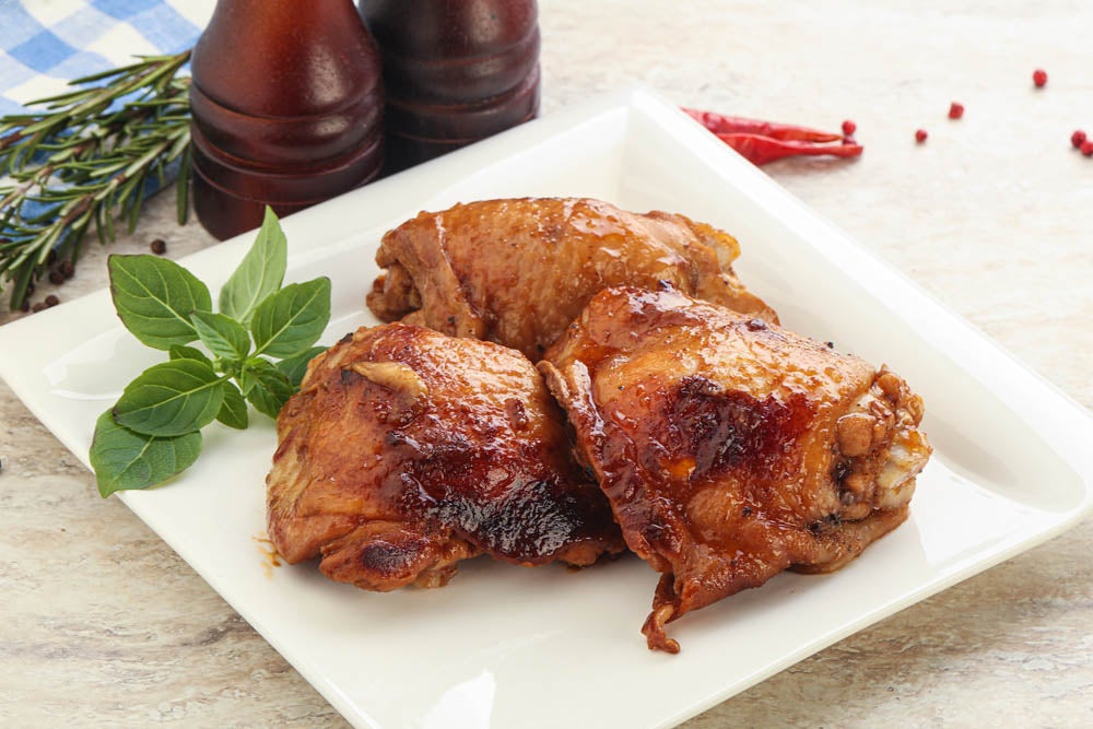 Tres muslos de pollo glaseados servidos en un plato blanco con una ramita de albahaca y condimentos de fondo.