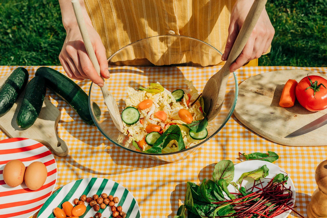 Woman mixing a salad on a table covered in ingredients outdoors.