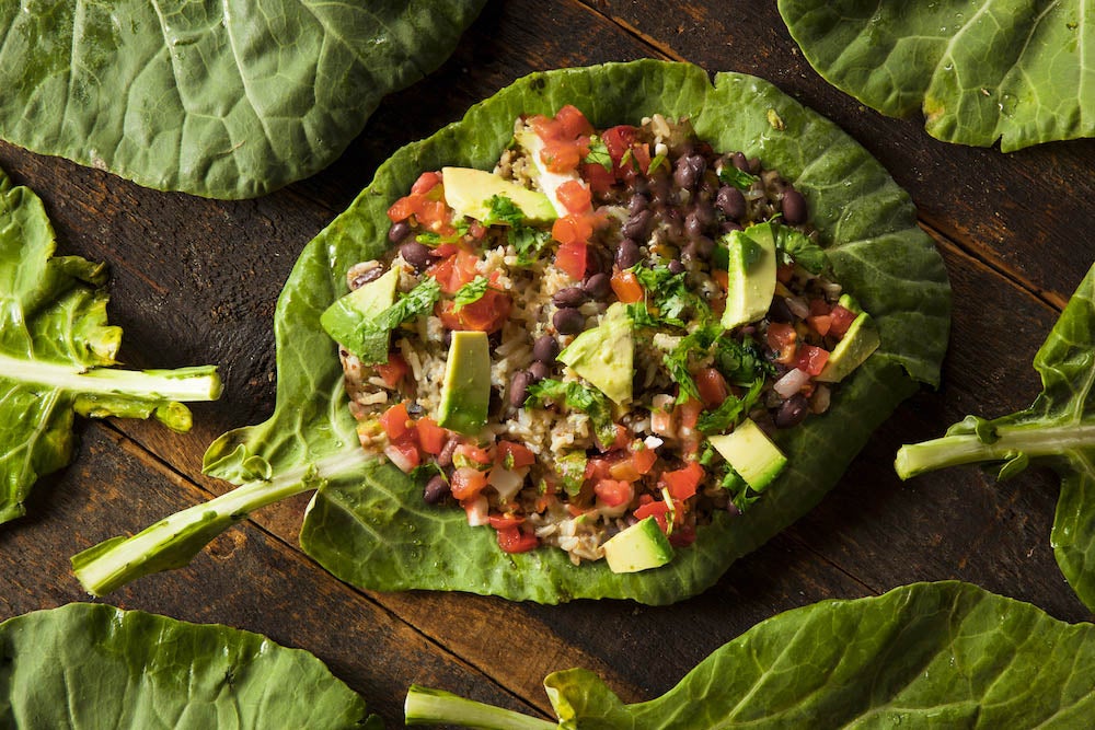 Hoja grande de collard rellena con una mezcla de frijoles negros, tomates en cubos, aguacate y hierbas, sobre una superficie de madera rodeada de más hojas de collard