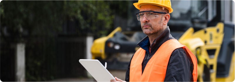 Construction worker in safety gear holding clipboard near road roller.