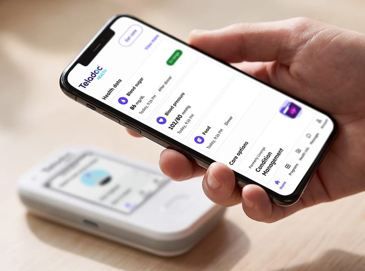Hand holding mobile phone with a blood glucose meter on a table in the background.