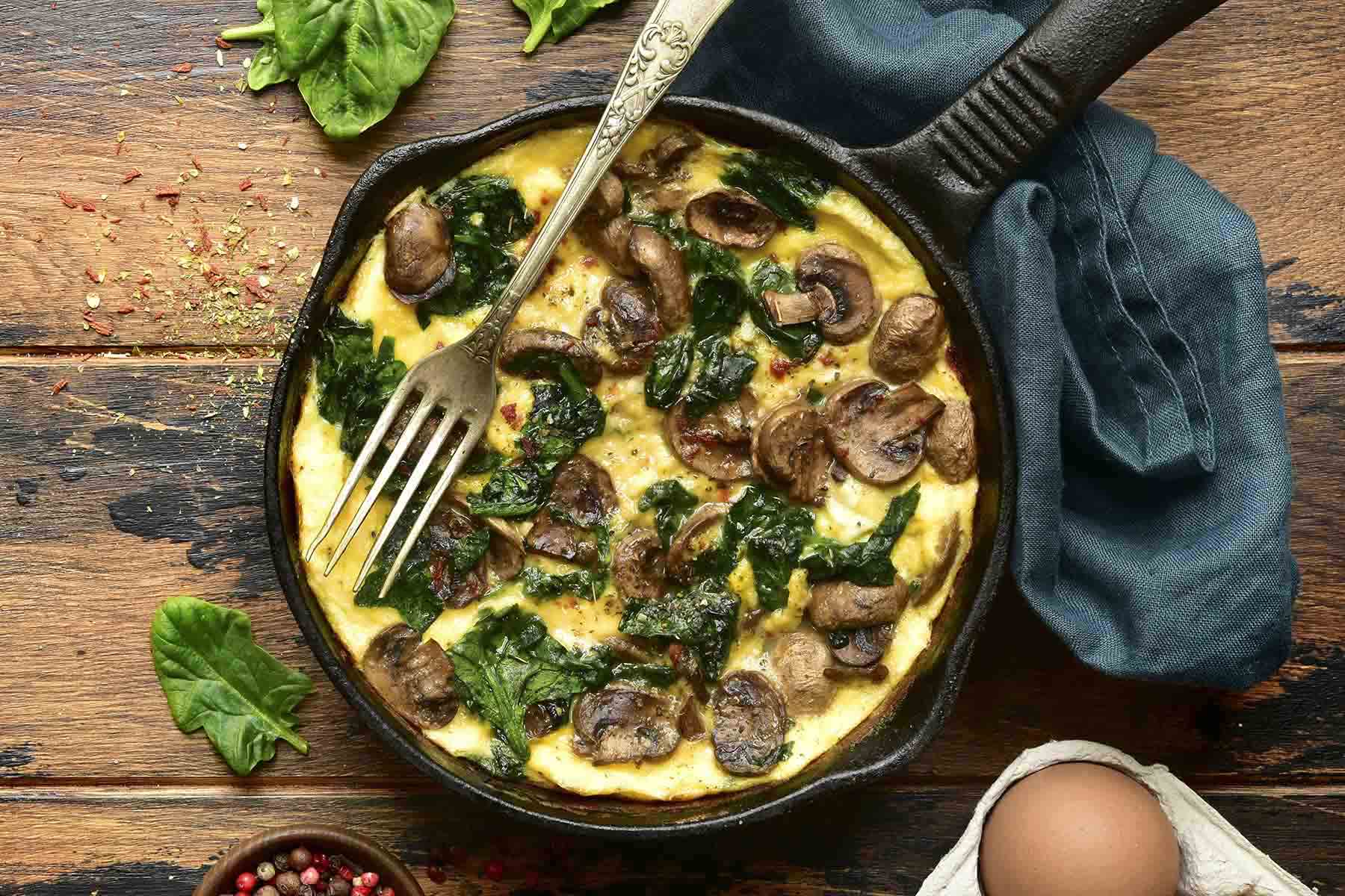  A skillet filled with a spinach and mushroom frittata, topped with a fork, surrounded by fresh spinach leaves and an egg on a wooden table.