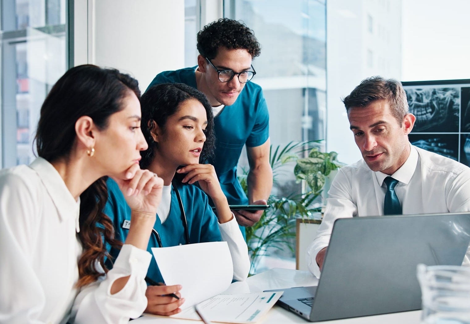 Clinical and administrative staff meeting around a laptop in a hospital conference room.