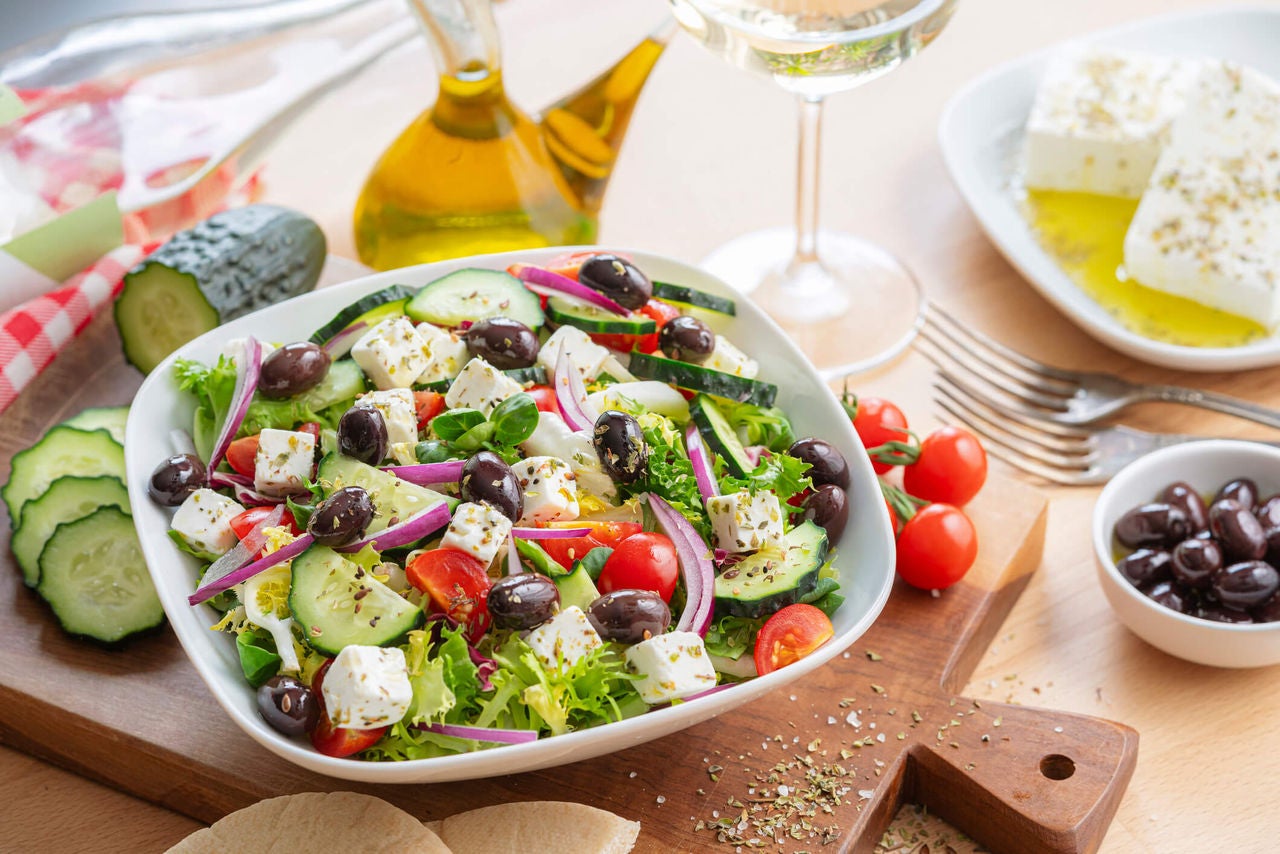 Bowl with olives, feta, cucumbers, and onions surrounded by indredients.