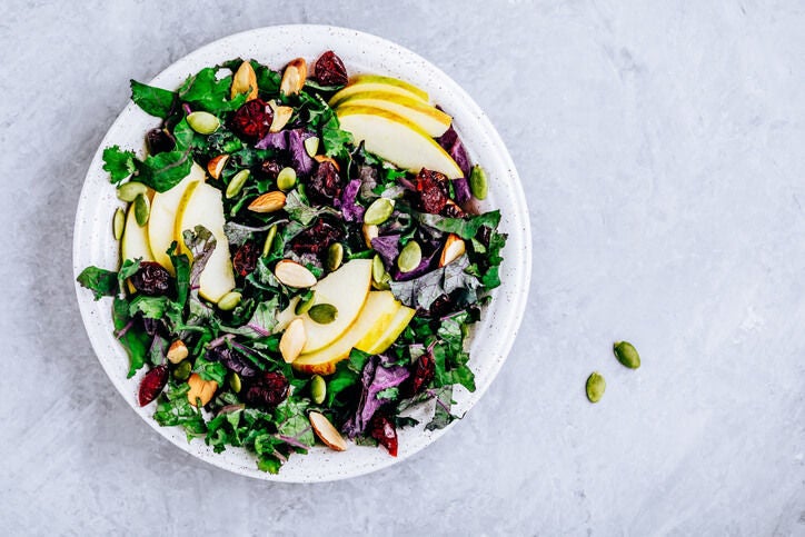 Bowl of salad with chopped kale, apple slices, seeds, almonds and dried fruit.