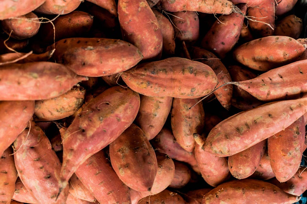 A pile of fresh sweet potatoes with reddish skin closely stacked together, filling the frame.