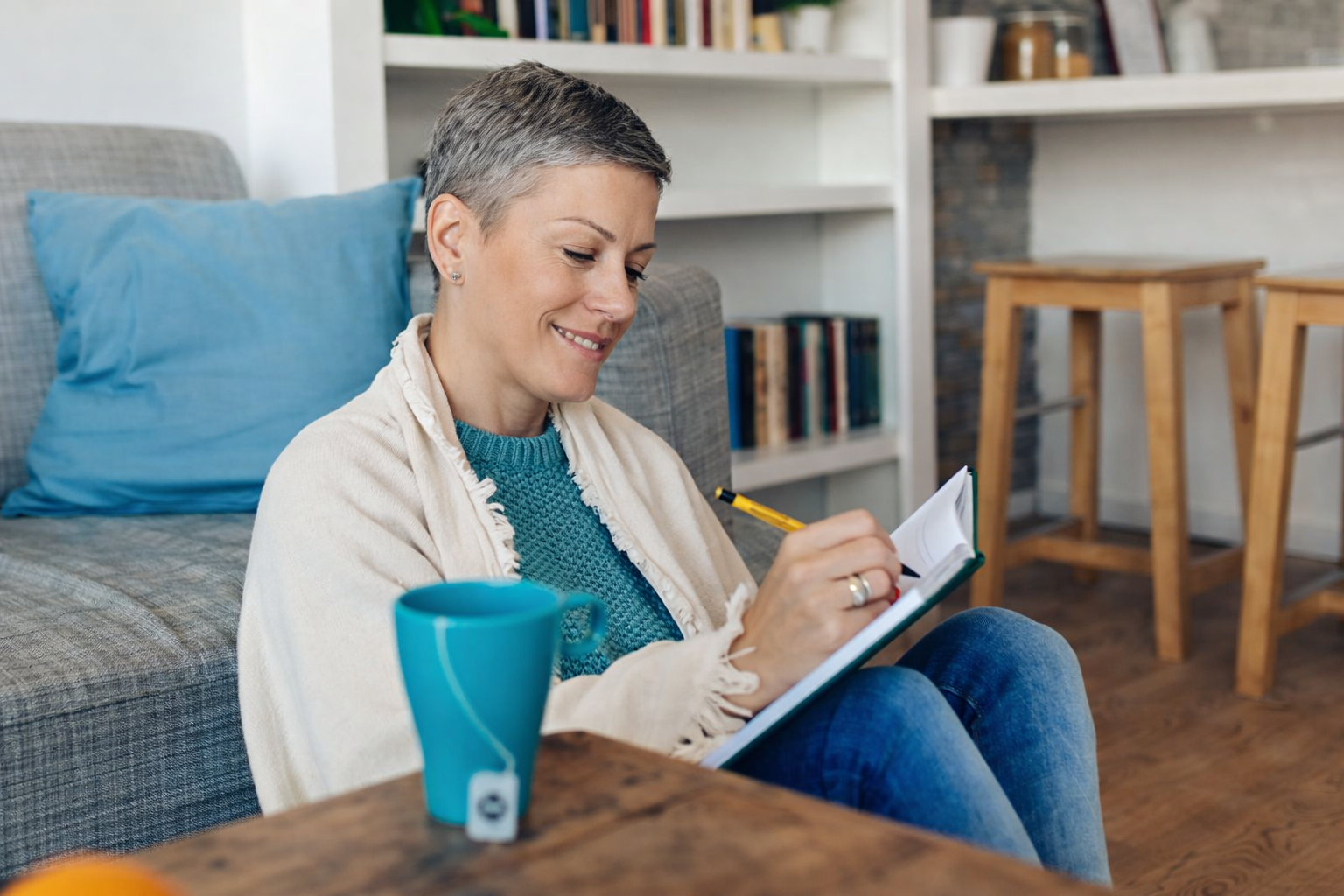 A smiling woman sitting on the floor, wrapped in a blanket, writing in a journal with a coffee cup beside her in a cozy living room setting.