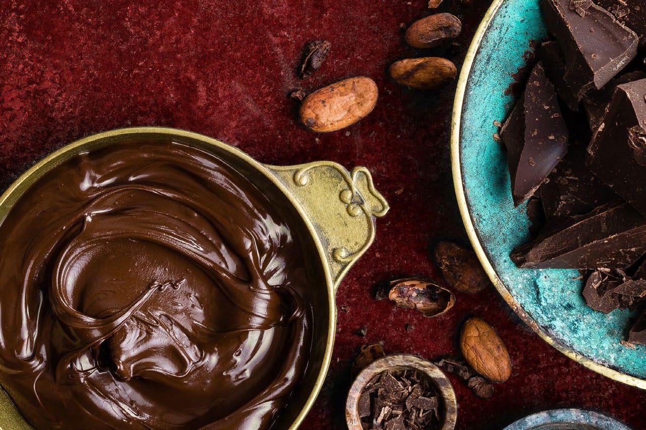 Rich melted chocolate swirls in a bowl on a burgundy surface, surrounded by cacao beans, chopped chocolate, and cacao nibs, evoking indulgence.