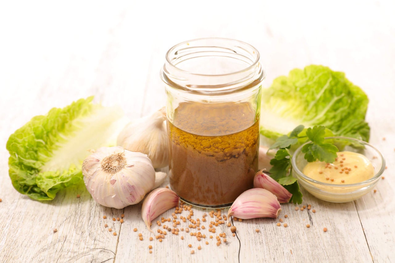 Jar of balsamic dressing next to cloves of garlic, mustard seeds and lettuce leaves.