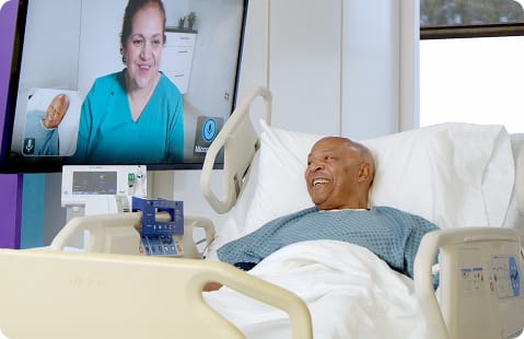 A patient in a hospital bed smiling while speaking with a virtual healthcare professional on a screen.