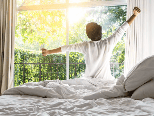 Person stretching while sitting on a bed, looking out a sunny window with trees outside.