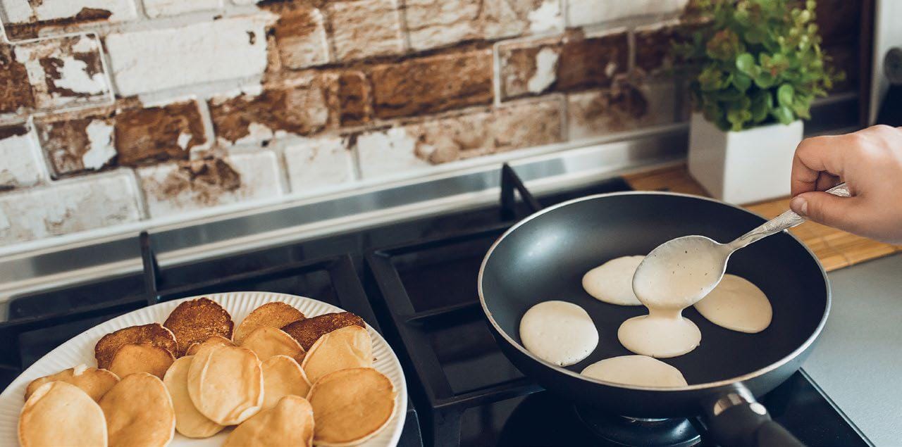Platter of pancakes on a gas stove next to a pan with cooking pancakes and a hand pouring pancake batter into the pan