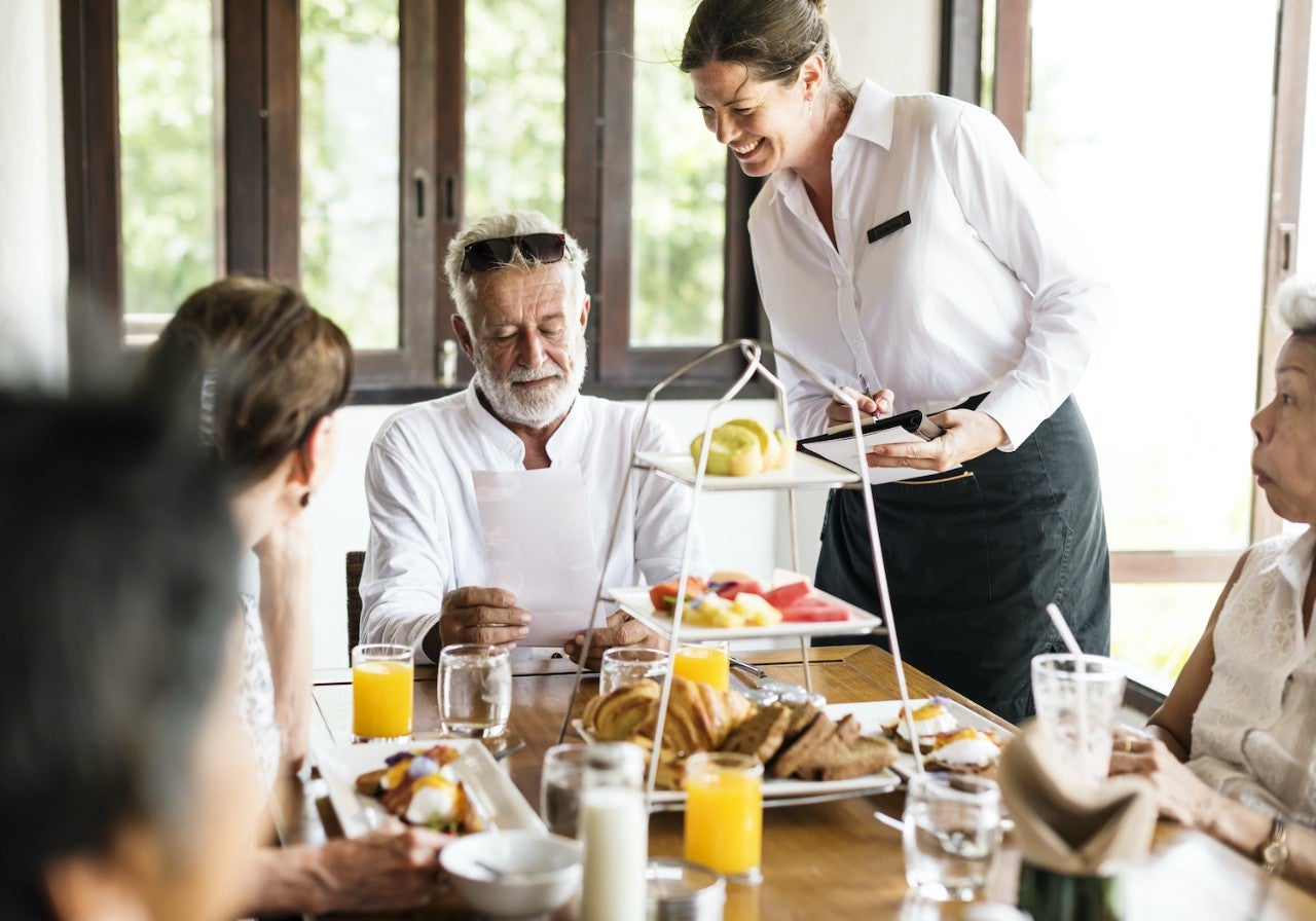 A server is presenting a dish to a group of diners at a table, where several people are enjoying a meal together in a bright and inviting restaurant setting.