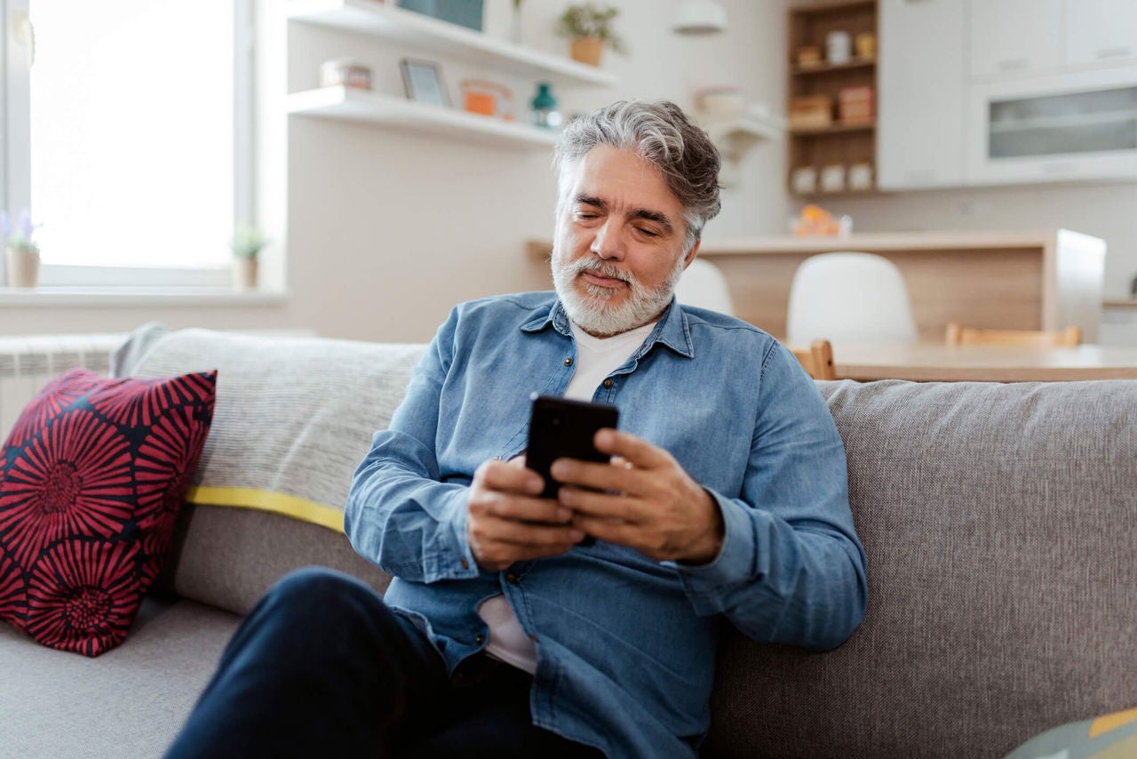 Man sitting on a couch in a living room holding and looking at a cell phone. 