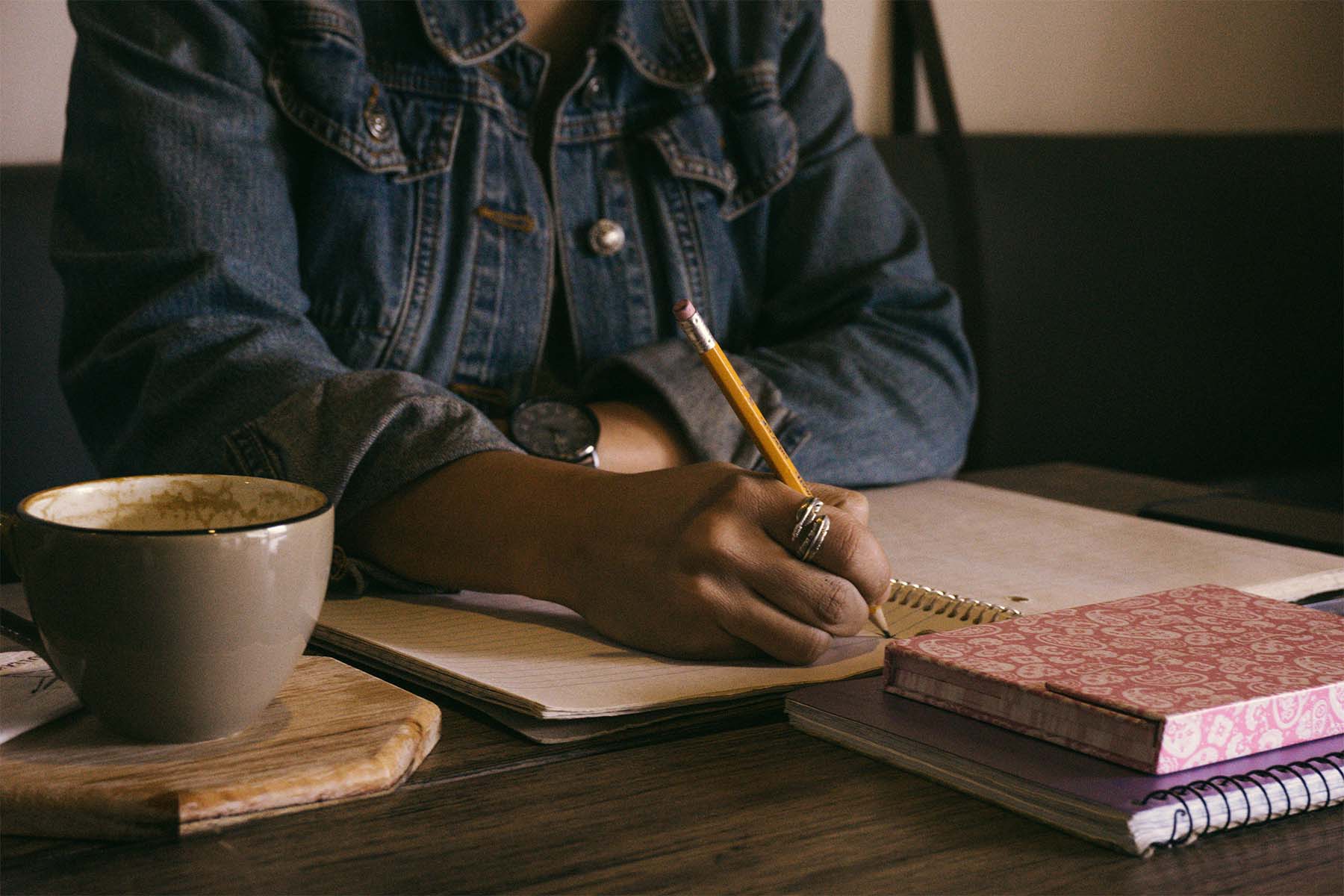 Person in a denim jacket writing with a pencil on a notebook. A mug and patterned notebook sit on the wooden table.