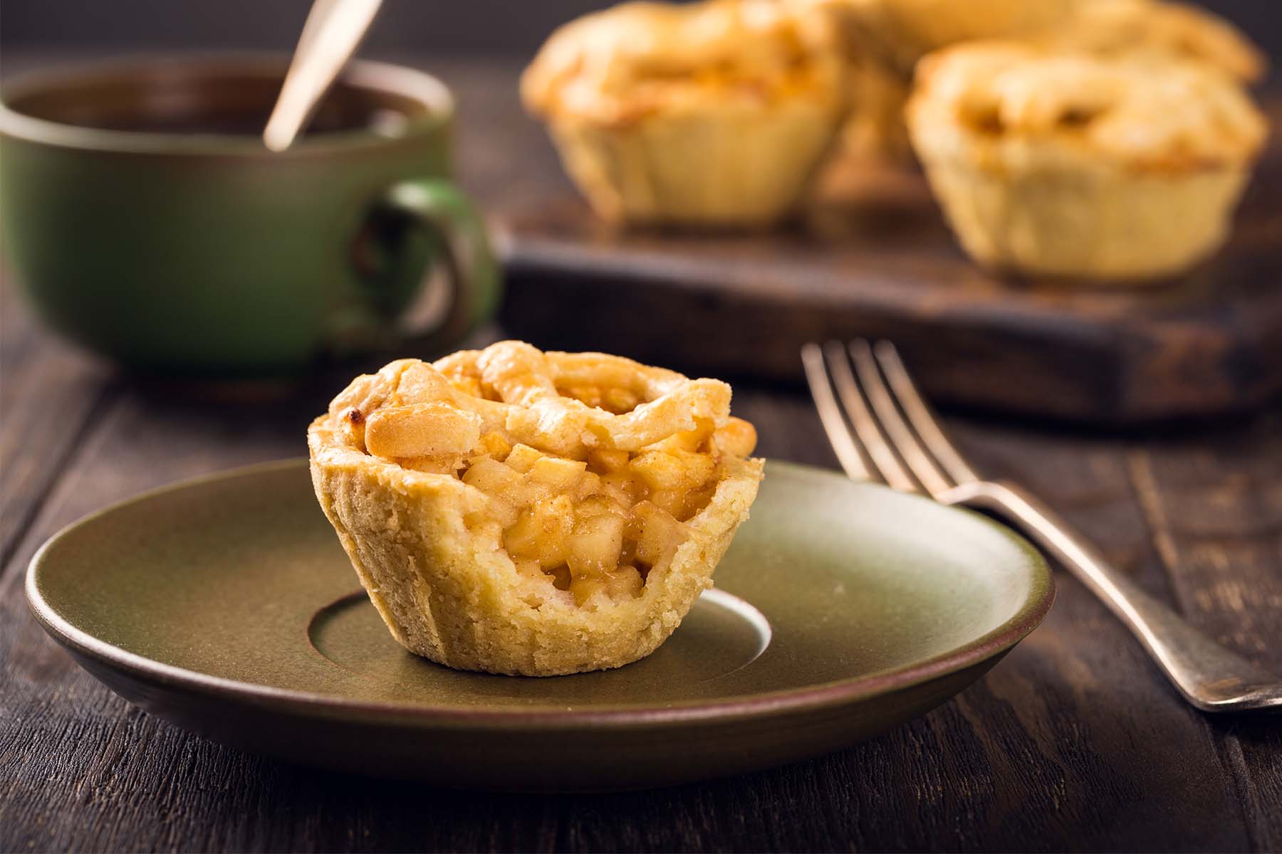 Mini apple tart with a flaky crust and caramelized apple filling, served on a green plate with a fork, with more tarts and a green mug in the background.