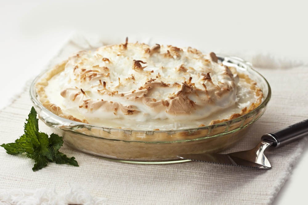 A lemon meringue pie with toasted meringue peaks in a glass pie dish, placed on a white cloth next to a pie server and mint leaves.