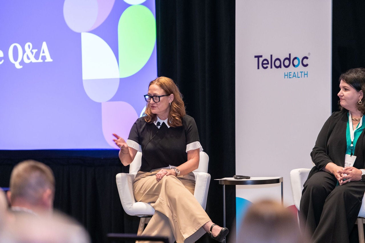 Two professionally dressed panelists sit on stage in white chairs having a discussion during Forum 2025. A panel behind them displays a Teladoc Health logo on a white background.